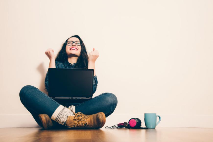 Young woman sittling on the floor with laptop in her lap, clenching her fists in a victory pose